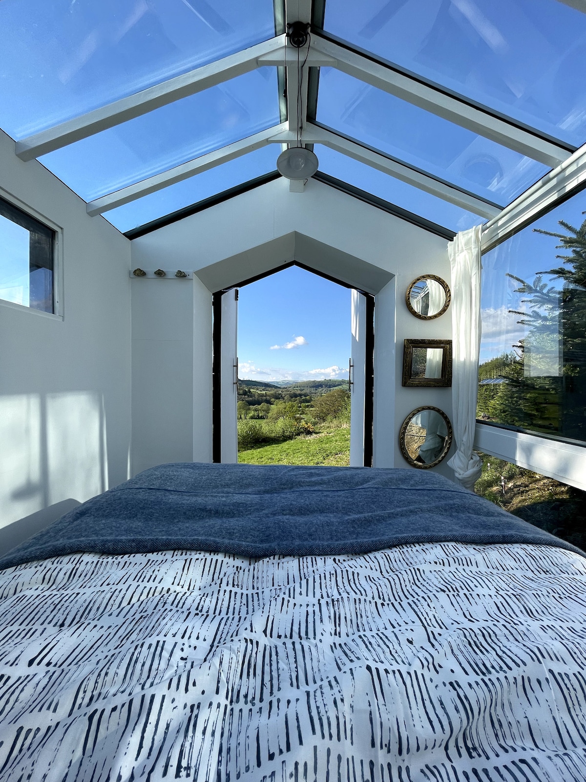 A bright sleeping area is showcased, featuring a spacious bed covered with a textured blanket. Large glass panels bring in natural light while framing stunning views of the surrounding landscape. Decorative mirrors are arranged on the wall, enhancing the spacious feel of the room.