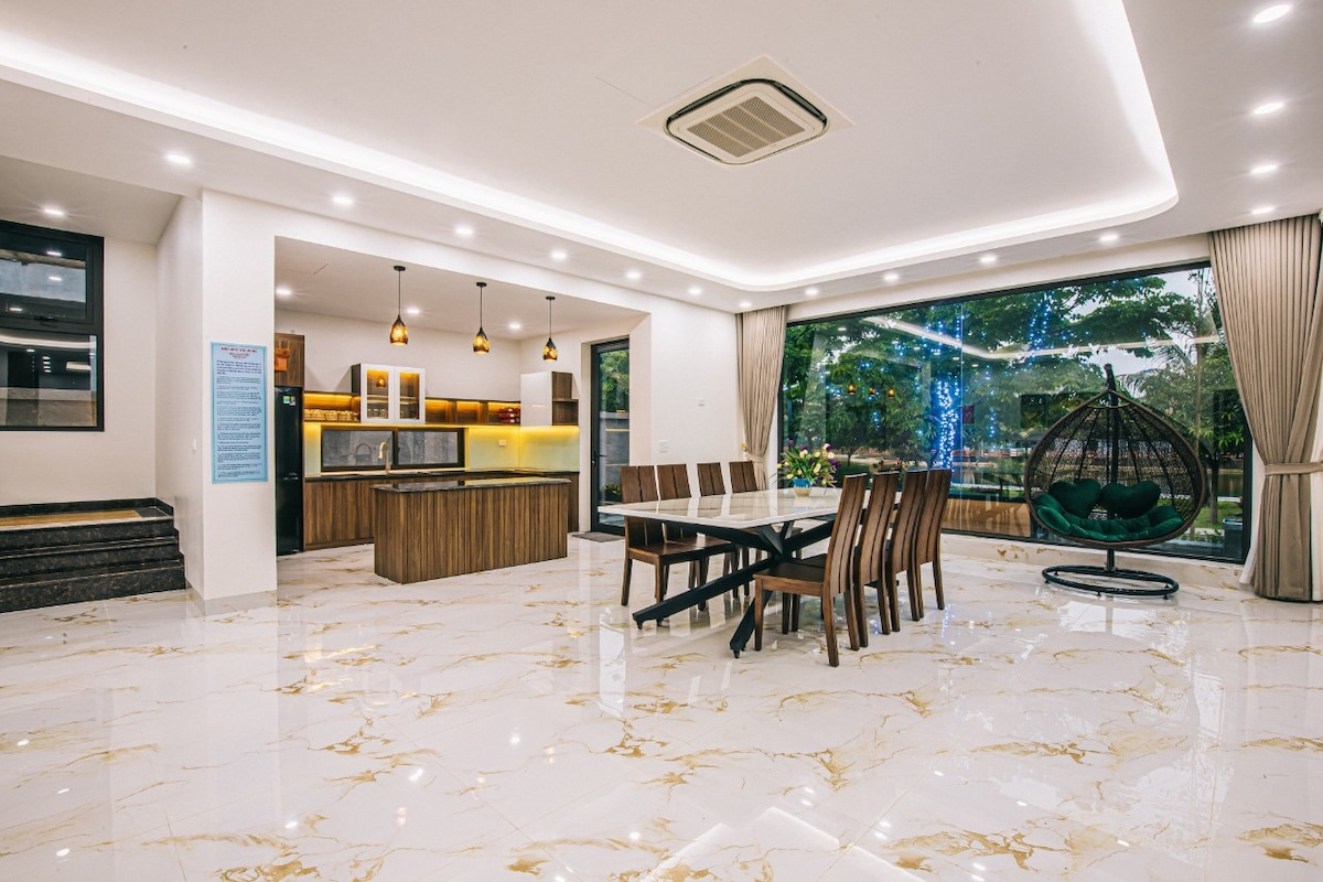 A spacious dining area is shown with a large table surrounded by multiple chairs. An open layout connects to a modern kitchen featuring sleek cabinets and a well-lit ambiance. Large windows provide views of the outdoor space, enhancing the bright and airy atmosphere.