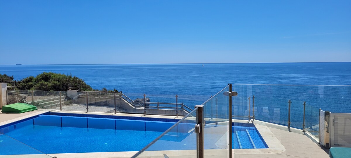 A clear view of a spacious infinity pool reflects the azure sky and sea. Glass railings surround the pool area, ensuring unobstructed vistas. Lush greenery is visible at the pool's edge, and the calm ocean stretches into the horizon.