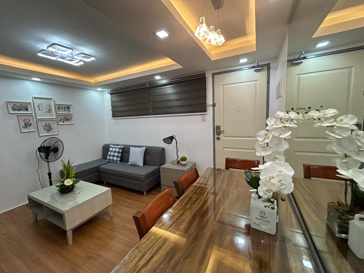 A cozy living area includes a gray sectional sofa and a coffee table adorned with a floral arrangement. A dining table with wooden chairs is positioned nearby. Wall art is displayed, and light fixtures contribute to a bright atmosphere. A fan is visible for air circulation.
