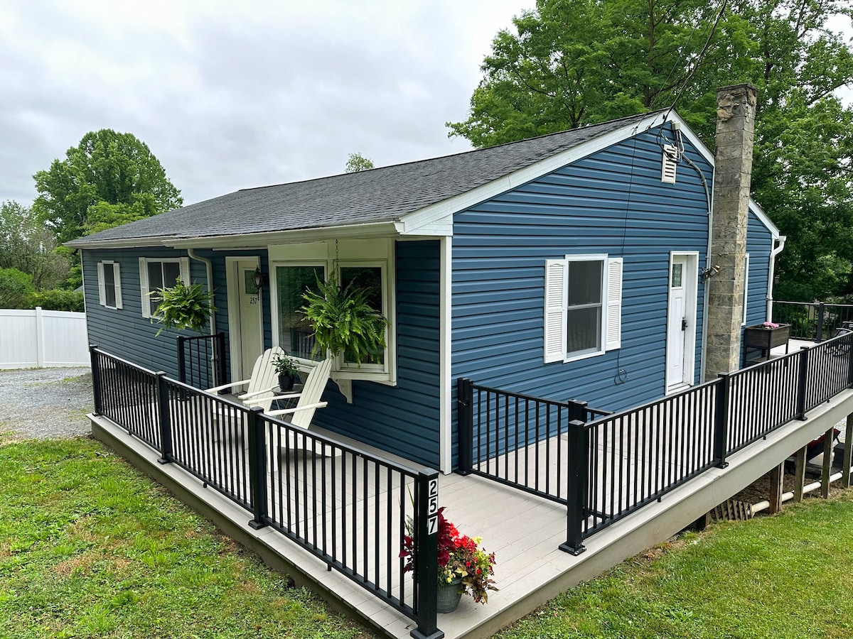A newly renovated cottage is set against a green backdrop, showcasing a blue exterior. The front porch features a light-colored rocking chair and is bordered by a black railing. Potted plants add a touch of color, while a stone chimney complements the home's design.