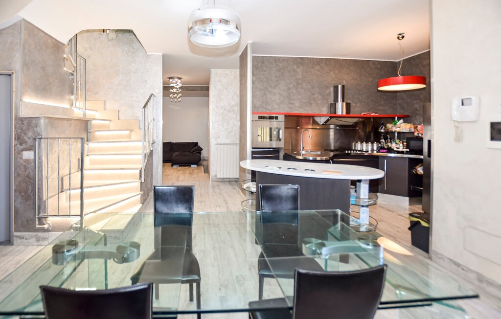 An open-concept kitchen and dining area features a glass dining table with four chairs. The kitchen is equipped with modern appliances and a large island. A staircase with illuminated steps leads to the upper level, while a comfortable seating area is visible in the background.