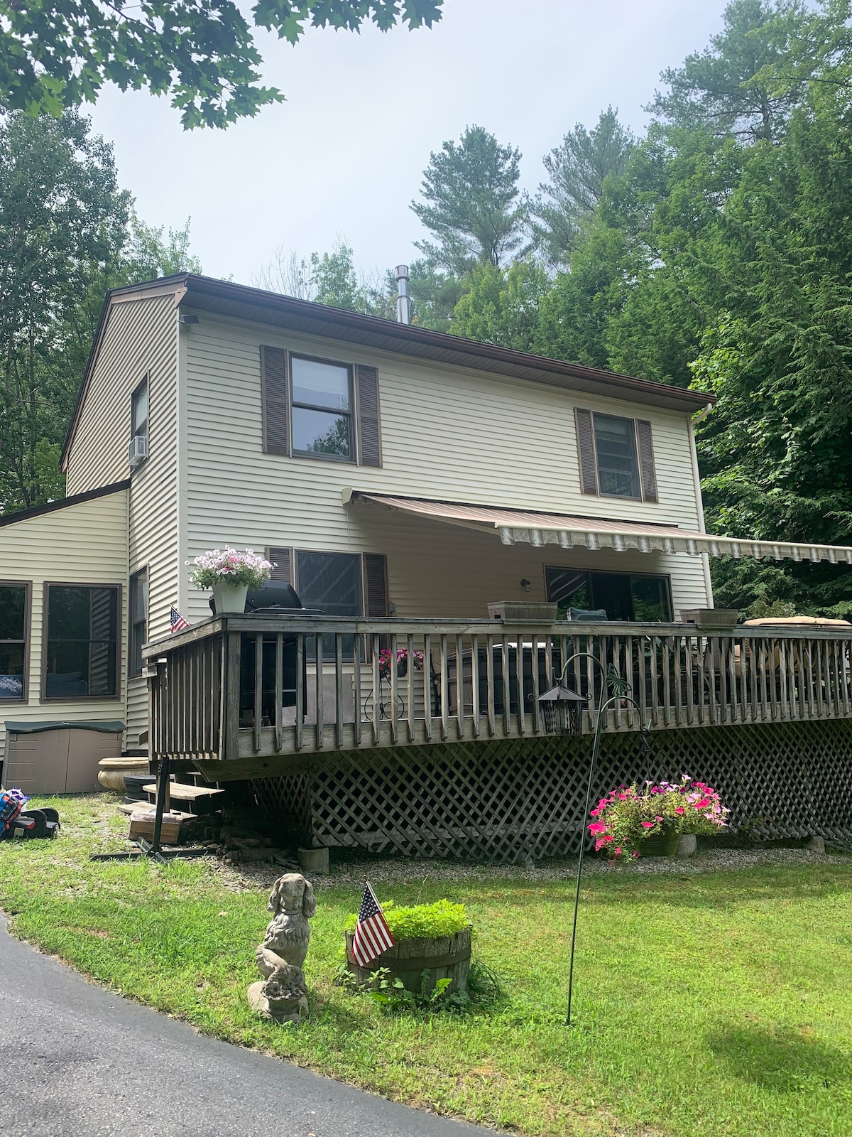 A two-story house is nestled among trees, offering a deck with an awning for shade. Flower pots adorn the entrance, and an American flag is visible in the foreground. The home's exterior features beige siding and brown window frames, creating a harmonious natural setting.