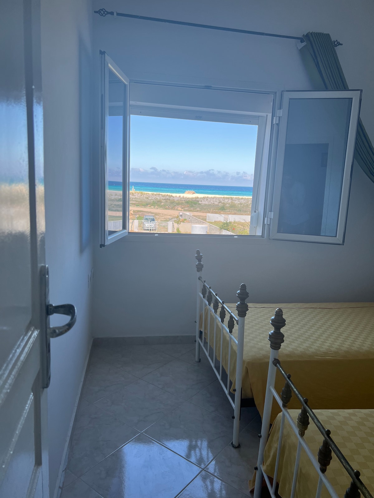 A bright bedroom features two twin beds with yellow bedspreads, positioned near a window. The window is ajar, offering a view of a coastal landscape with water in the background. Soft natural light fills the space, highlighting the room's simplicity.