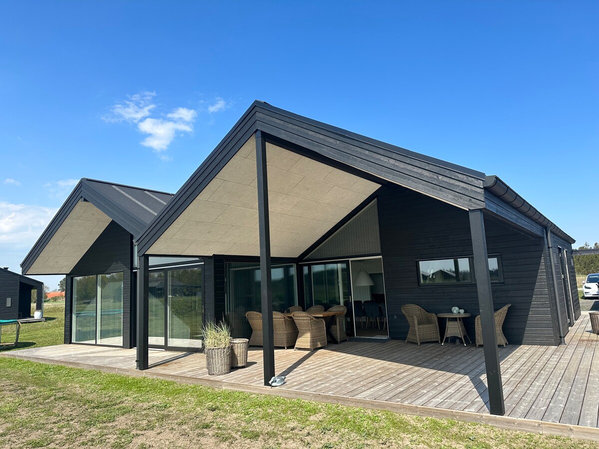 A modern summer house features a distinctive roof design and large glass doors, allowing natural light to fill the interior. A spacious wooden deck is lined with seating areas for outdoor relaxation. Green grass surrounds the home, enhancing the natural setting.