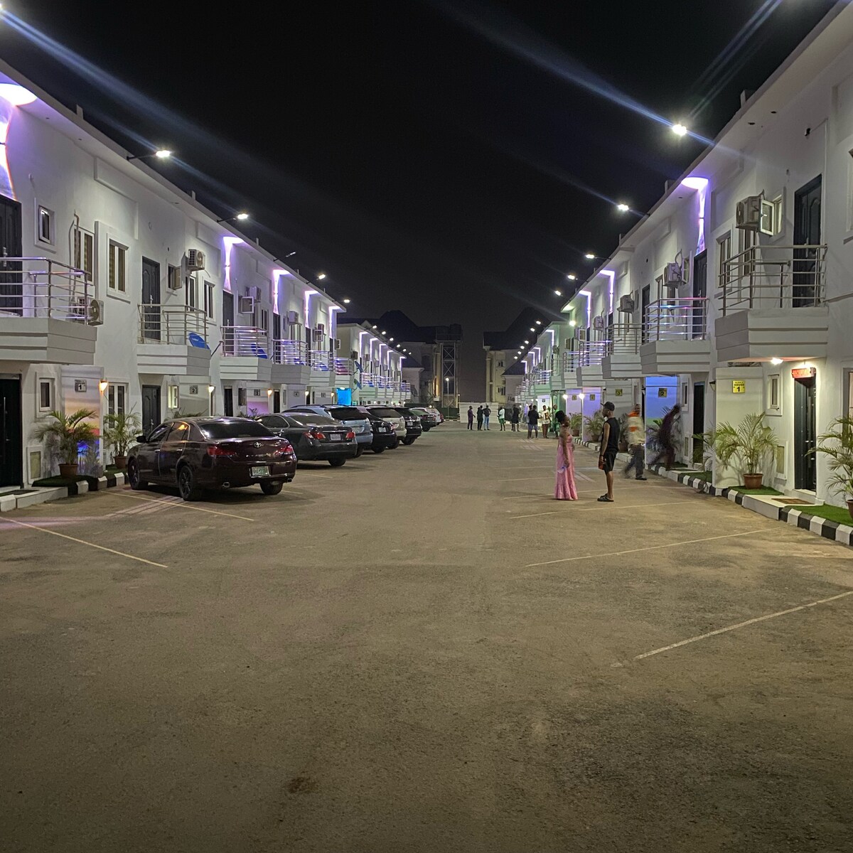 The image captures a well-lit courtyard at night, featuring two rows of modern apartments adorned with soft purple lighting. Vehicles are parked along the paved pathway, with guests mingling in the open area, creating a lively yet welcoming atmosphere.