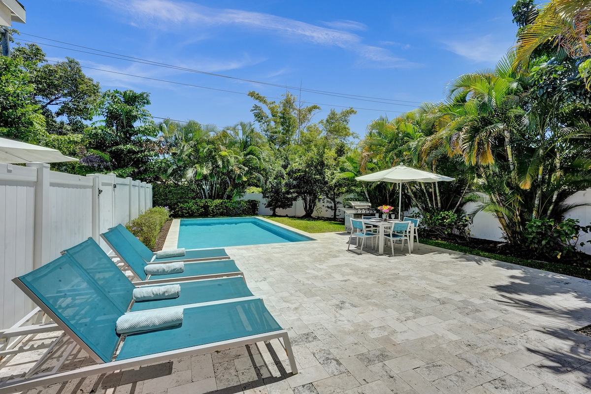 A private backyard features a swimming pool surrounded by lush greenery. Lounge chairs are positioned beside the pool, enhancing relaxation opportunities. An outdoor dining table with umbrellas invites al fresco meals, and a patio area with stone flooring provides ample space for guests to enjoy the serene environment.