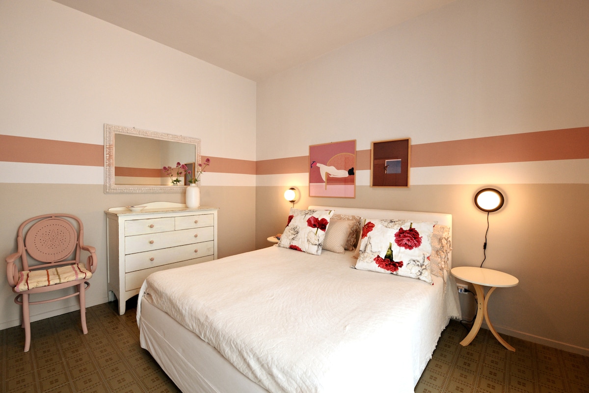 A serene bedroom is presented with a spacious bed adorned with floral cushions. Soft color blocking in shades of white and pink creates a calming effect. A stylish dresser and a small chair add charm, while wall art enhances the modern aesthetic.