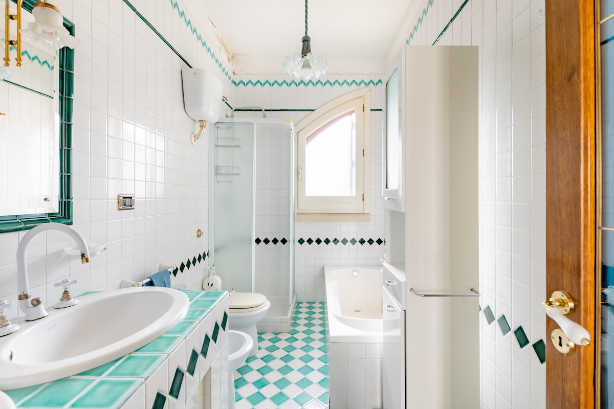 A well-appointed bathroom features white tiled walls and a distinctive green and white checkered floor. A glass shower is positioned next to a window, allowing natural light. A large bathtub and dual sinks contribute to the spaciousness, with modern fixtures completing the inviting atmosphere.
