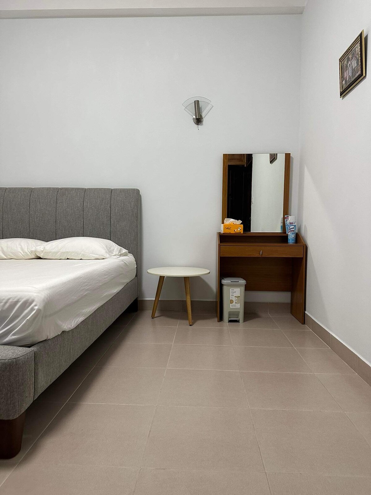 A room with a gray upholstered double bed and a white bedding set is depicted. A small white side table stands beside the bed. A wooden makeup table with a mirror is positioned across from the bed, with various toiletries neatly arranged on top.