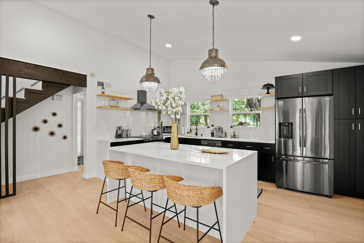 A modern kitchen showcases sleek black cabinets and stainless steel appliances. A large island with seating for three is accented by decorative light fixtures overhead. Natural light streams through the windows, highlighting the minimalist design and wooden flooring.