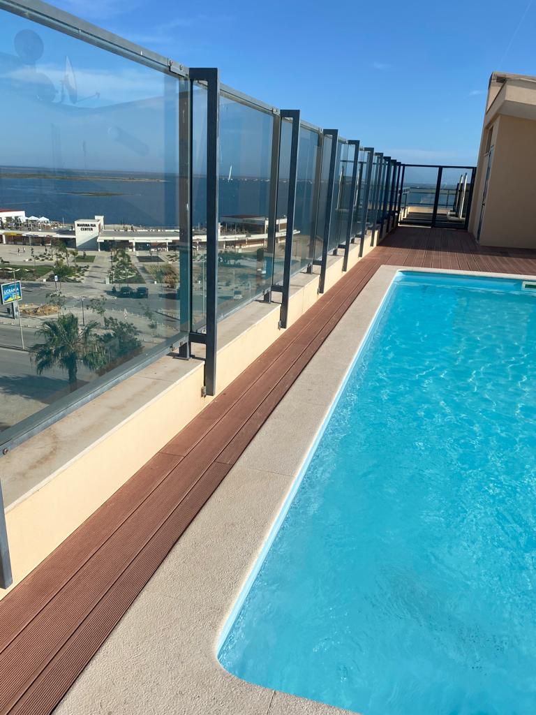 A rooftop area features a clear swimming pool bordered by a wooden deck, with panoramic views overlooking the marina and the sea. Glass railings provide an unobstructed sight of the surroundings, contributing to an open and airy feel.