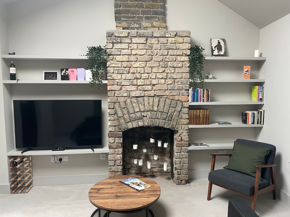 A living room features a stone fireplace with decorative candles arranged within. Light grey walls complement wooden shelves, which are adorned with books and decorative items. A comfortable armchair and a round coffee table provide inviting seating, while a flat-screen TV is positioned on the wall.