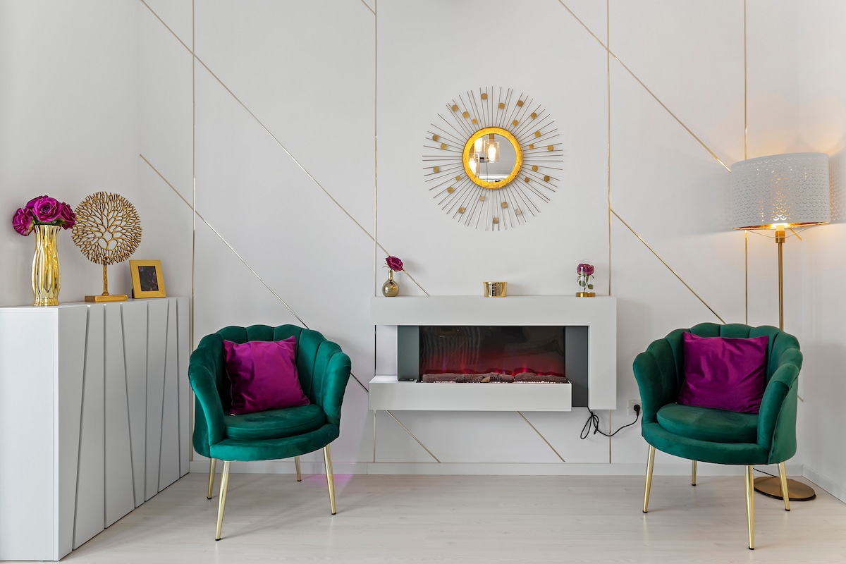 A stylish seating area is highlighted by two green velvet chairs with gold legs, facing a modern electric fireplace. A sunburst mirror adorns the wall above the fireplace, which is framed by minimalist gold accents. Decorative vases with flowers and framed photos add a personal touch.
