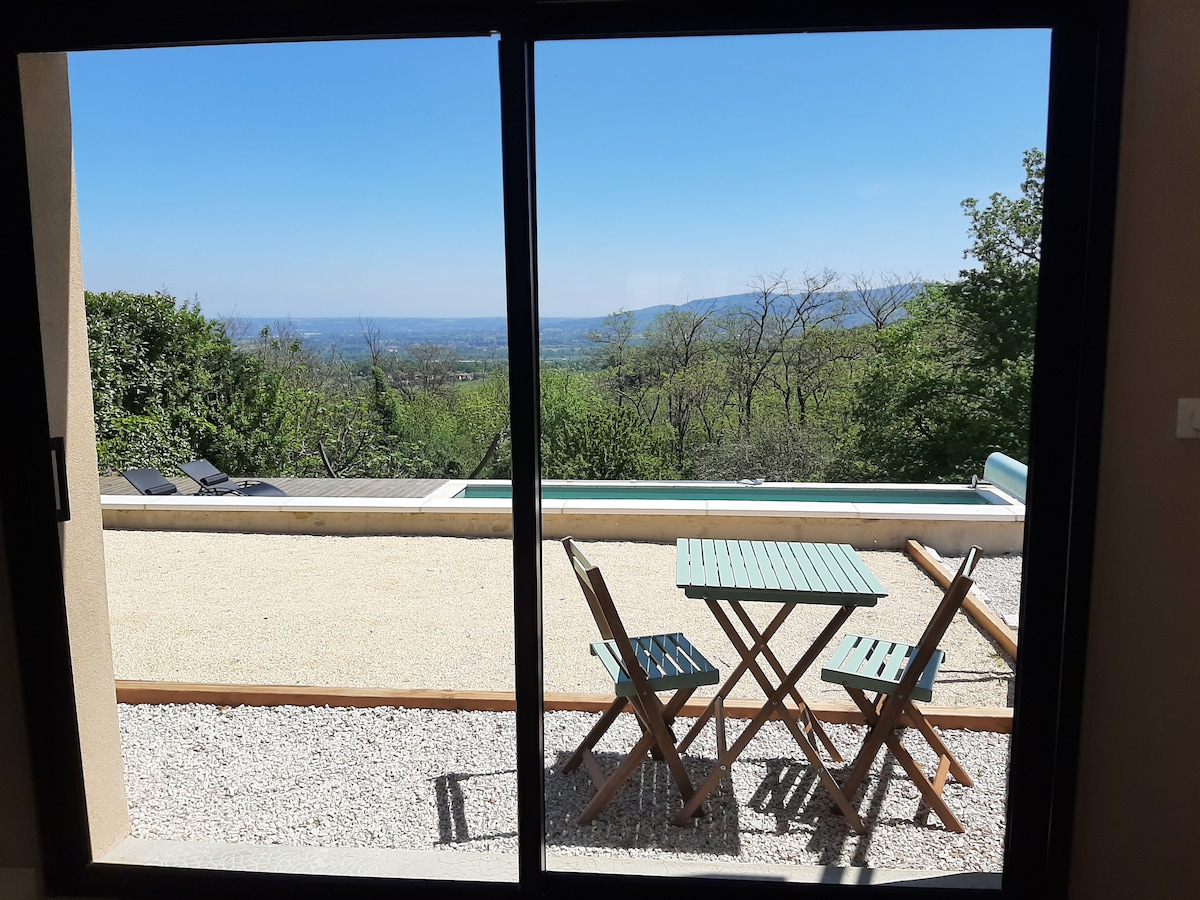 A view of the outdoor area from the interior through a large glass door. A wooden table with two folding chairs is visible on a gravel surface, with a swimming pool and a panorama of the surrounding greenery extending in the background.