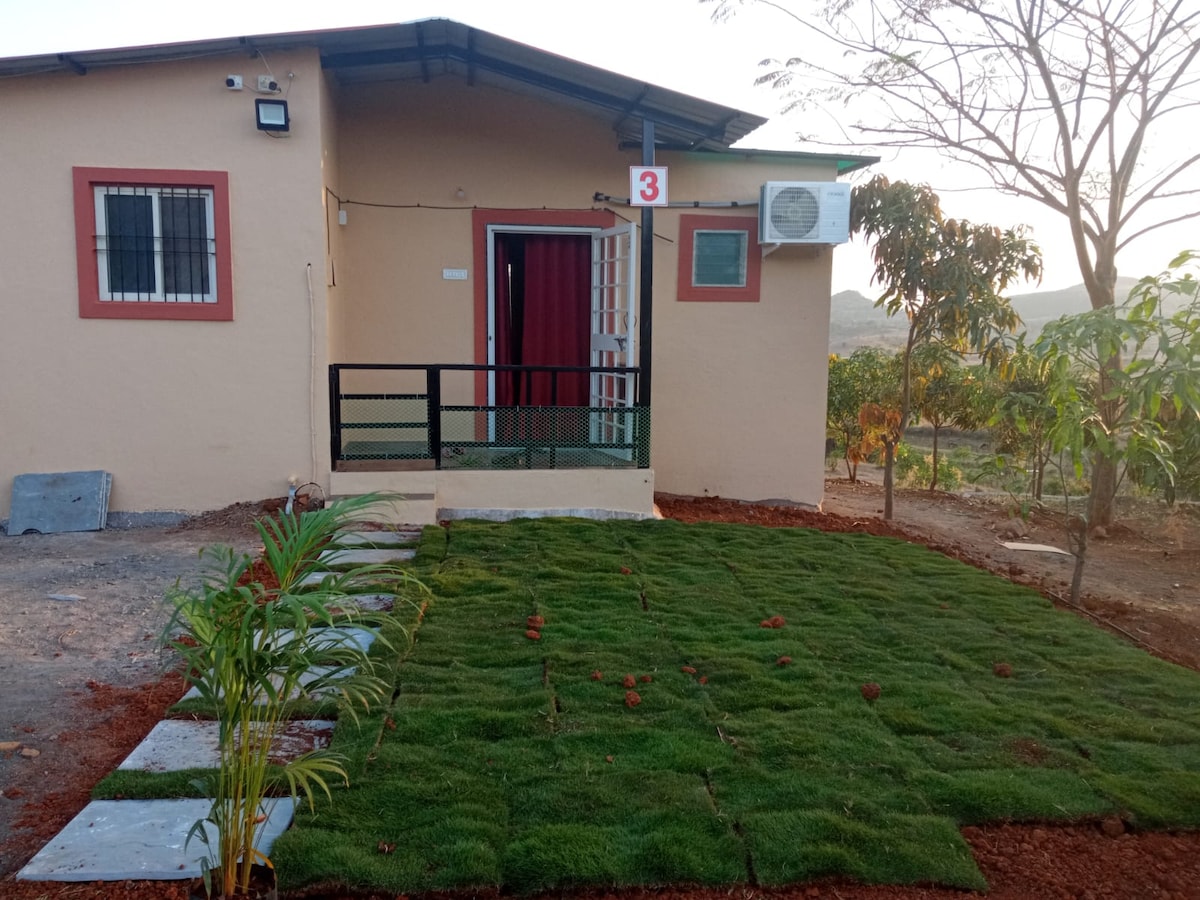 The exterior of the cottage is visible, featuring a small entryway with a designated door number. A green lawn made of grass serves as a welcoming pathway to the entrance. A window and an air conditioning unit are positioned next to the door, providing comfort. Surrounding trees can be seen in the background.