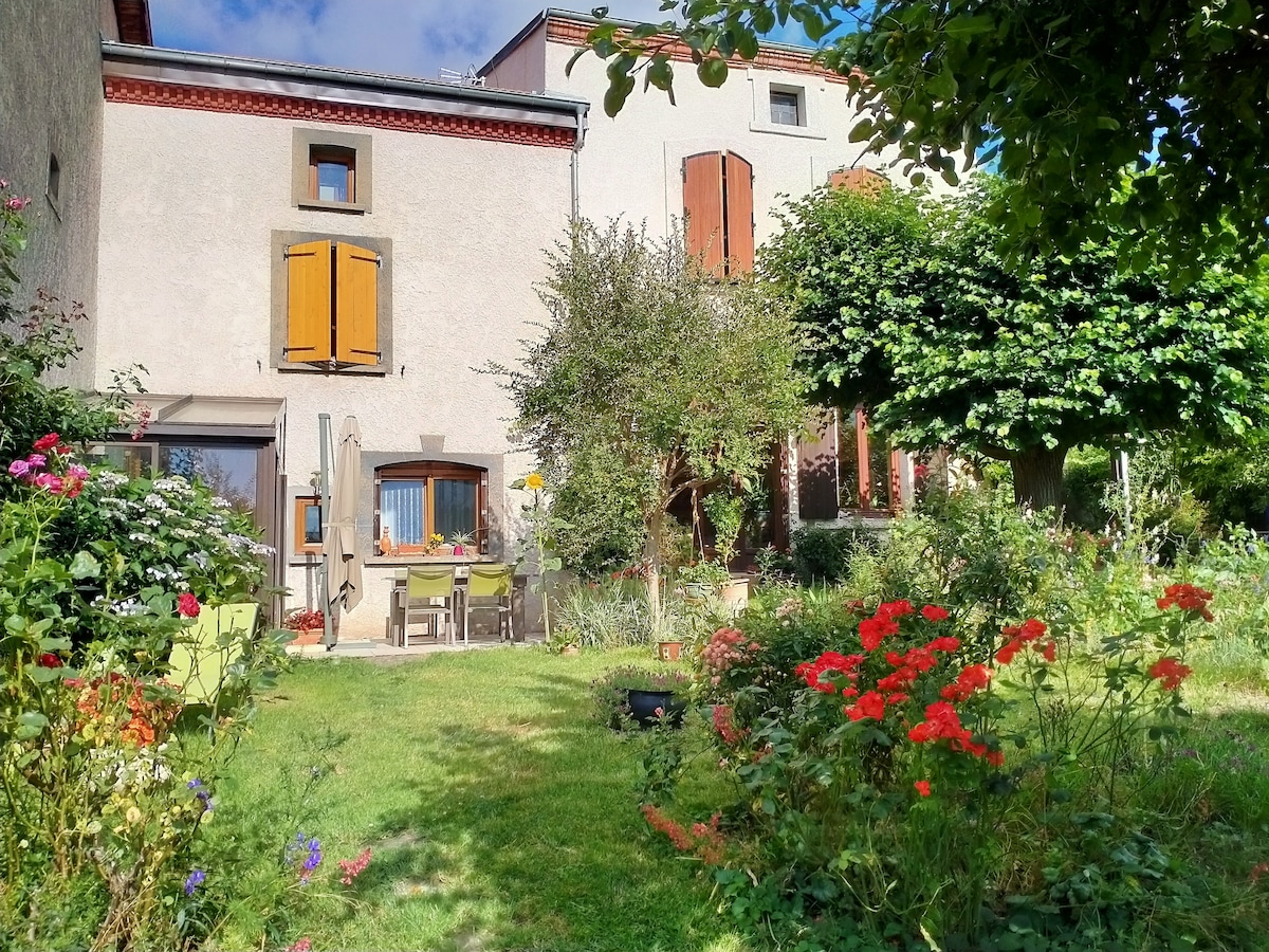 The exterior of the accommodation reveals a charming garden filled with colorful flowers and greenery. A patio area features a table and chairs under the shade of a large tree. The building showcases large windows with wooden shutters and a well-maintained facade.