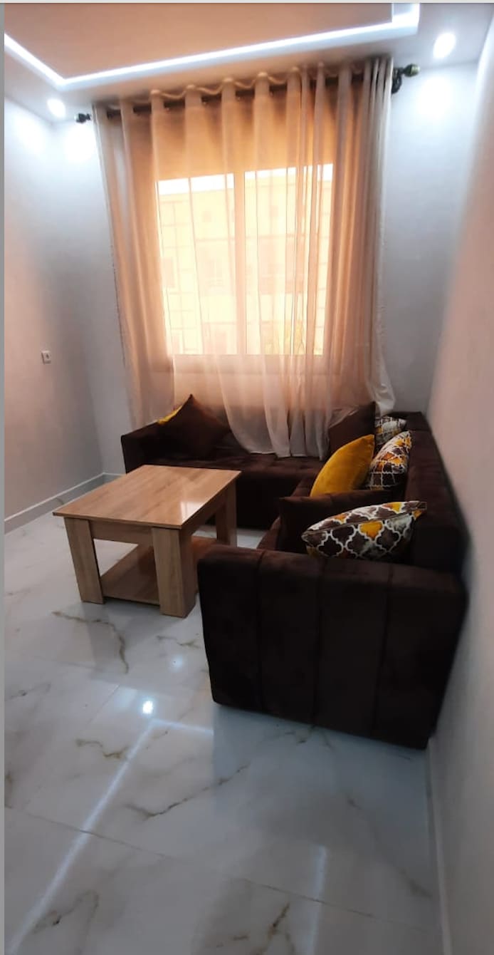 A living area is presented with a comfortable brown sectional sofa adorned with colorful throw pillows. A light wooden coffee table is positioned in front of the seating area. Natural light filters through sheer curtains, creating a bright and airy ambiance.