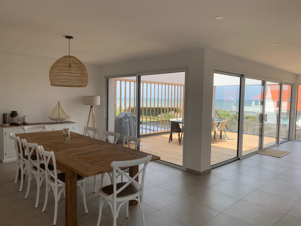 A spacious dining area features a large wooden table surrounded by white chairs. Expansive glass doors open to a wooden terrace, offering views of the sea. Natural light fills the room, highlighting the neutral décor and a decorative woven light fixture above the dining table.