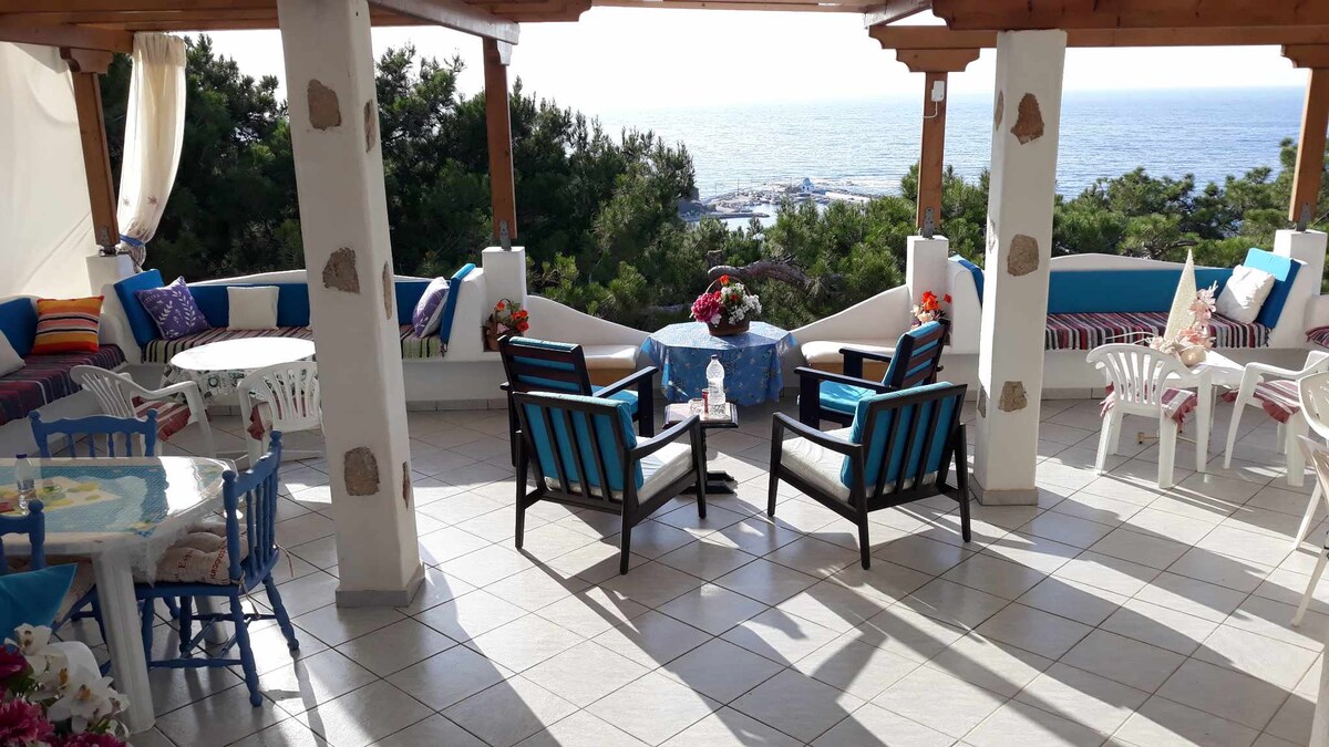 An open-air terrace features comfortable seating arrangements with blue and white accents. Tables are set, accompanied by vibrant cushions and decorative flowers. The sea view is visible in the background, enhancing the serene atmosphere of the space.