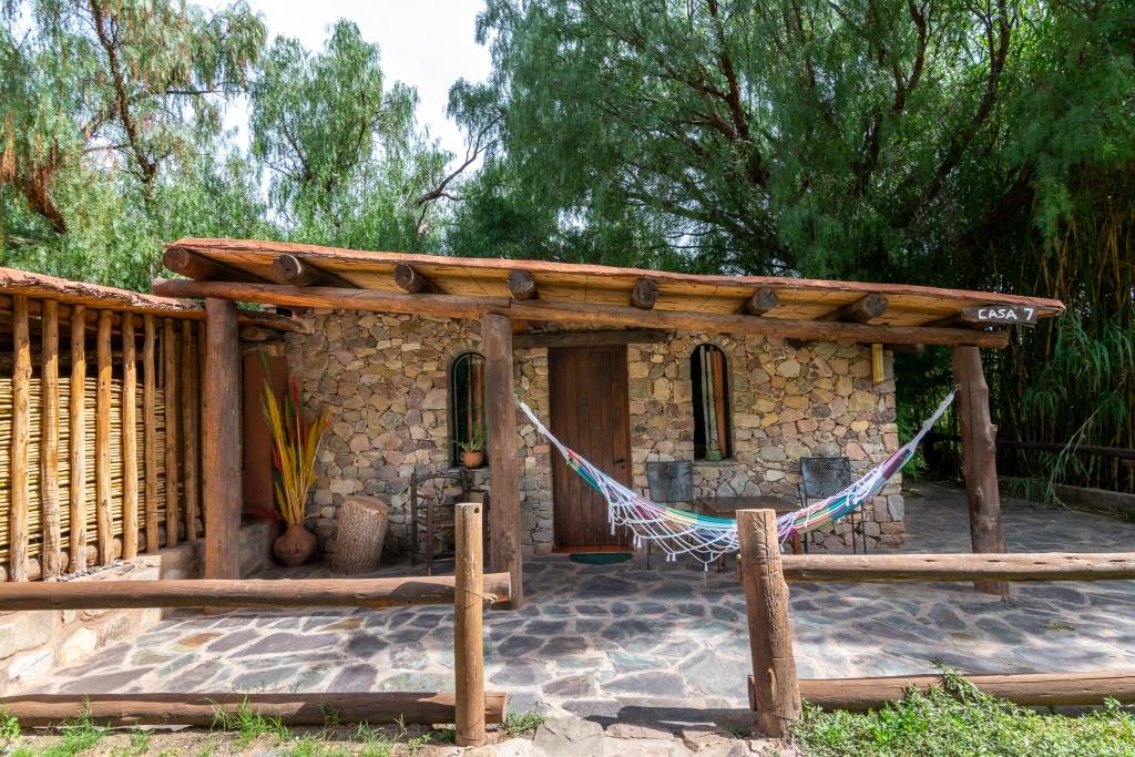 A rustic stone cottage features a thatched roof and a welcoming entrance. A colorful hammock is displayed outside, offering a comfortable spot to relax. Surrounding greenery enhances the outdoor space, complemented by stone pathways and wooden posts marking the area.