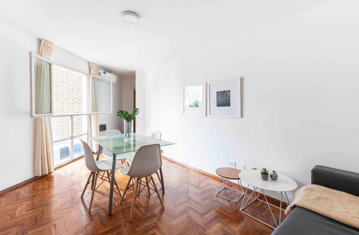 A bright living area is presented with a glass dining table surrounded by four chairs. Natural light pours in through open windows, illuminating the wooden floor. A gray sofa and two small side tables are situated nearby, creating an inviting space for relaxation and dining.