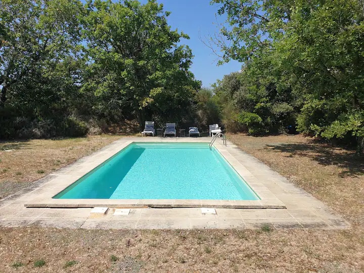 A private swimming pool is surrounded by natural landscaping, offering a serene setting. The clear water reflects sunlight and is bordered by a stone terrace. Several sun loungers are positioned nearby, inviting relaxation in the outdoor space.