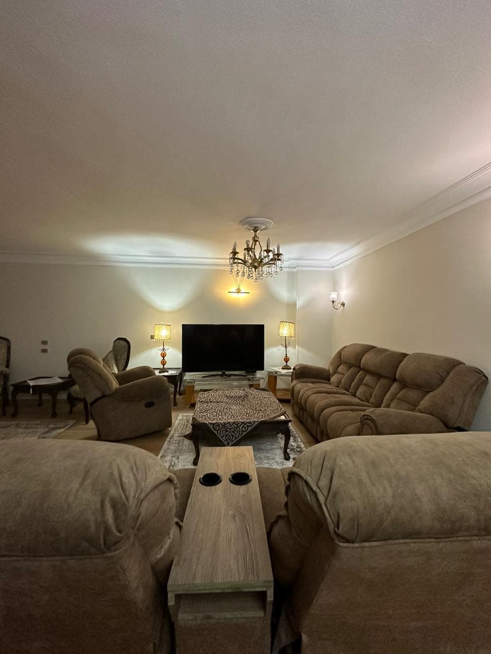 A cozy living area is presented, featuring plush seating arranged around a decorative coffee table. A large television is positioned on a cabinet, and two elegant lamps provide soft lighting. A chandelier adds a touch of sophistication to the space.