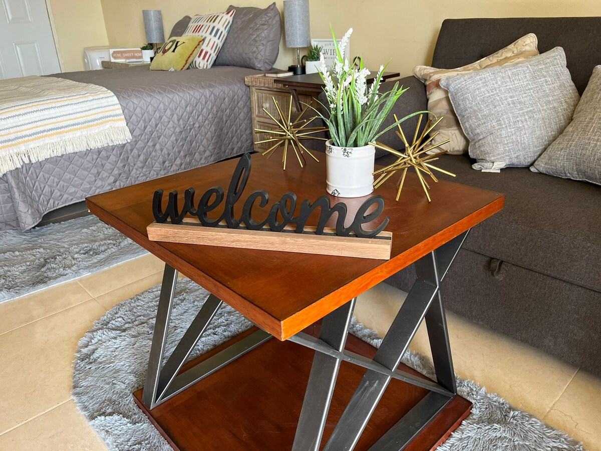 A wooden coffee table with a modern design is positioned in front of a dark sofa. A decorative 'welcome' sign rests atop the table, complemented by a potted plant and gold decorative accents. Soft area rugs are placed beneath the furniture for added comfort.