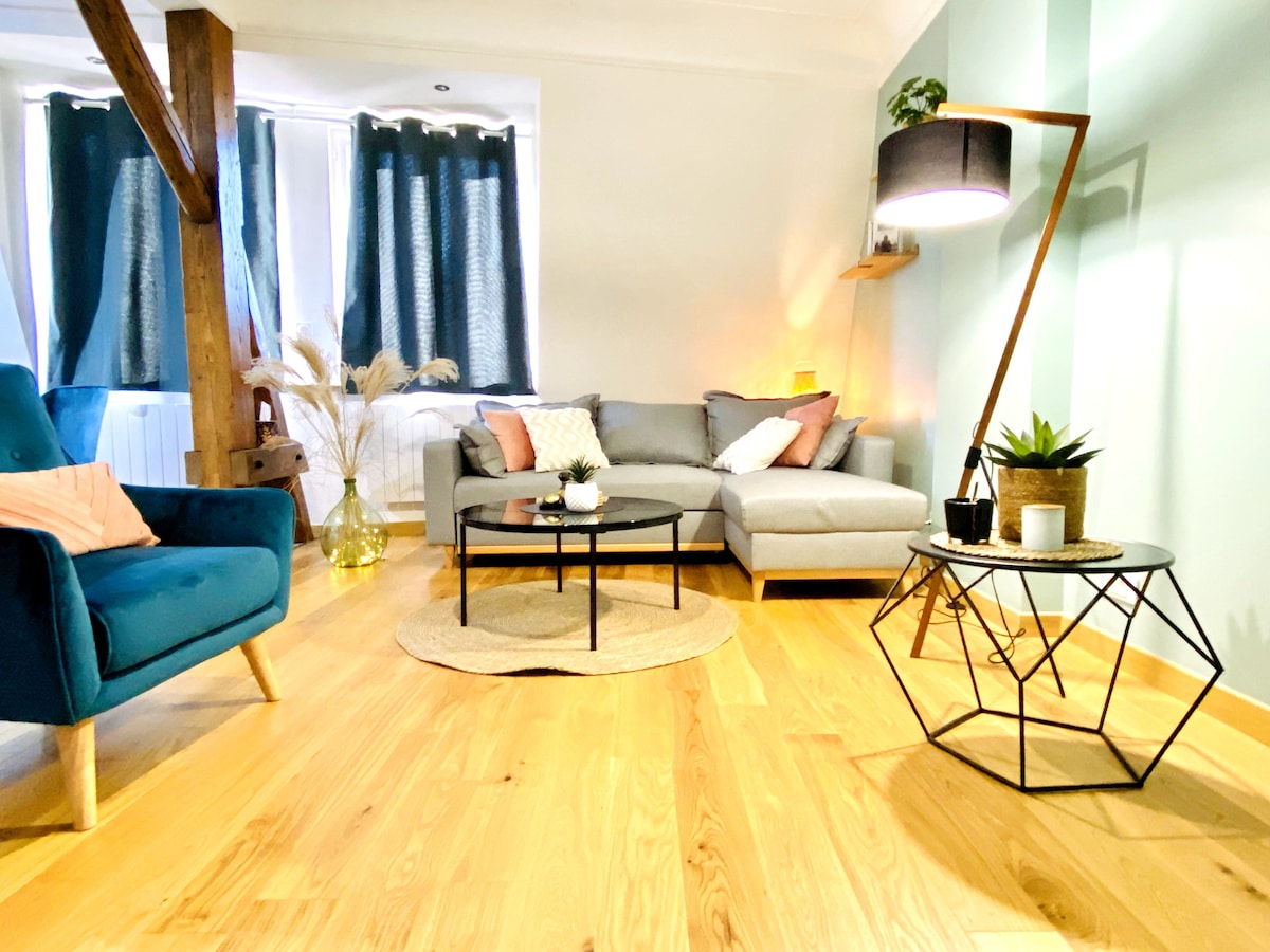 A bright living area features a comfortable gray sectional sofa, accented by decorative pillows. A sleek coffee table stands on warm wood flooring, complemented by a stylish armchair in teal. Large windows with dark curtains allow natural light to fill the space.