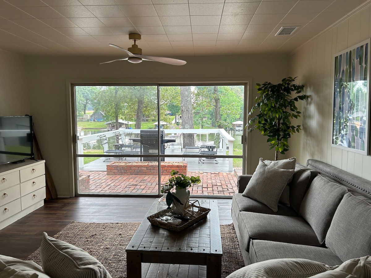 A bright living area features a gray sofa and a light wood coffee table, adorned with a decorative tray and a vase of flowers. Large sliding glass doors provide a view of the outdoor deck, framed by greenery and natural light.