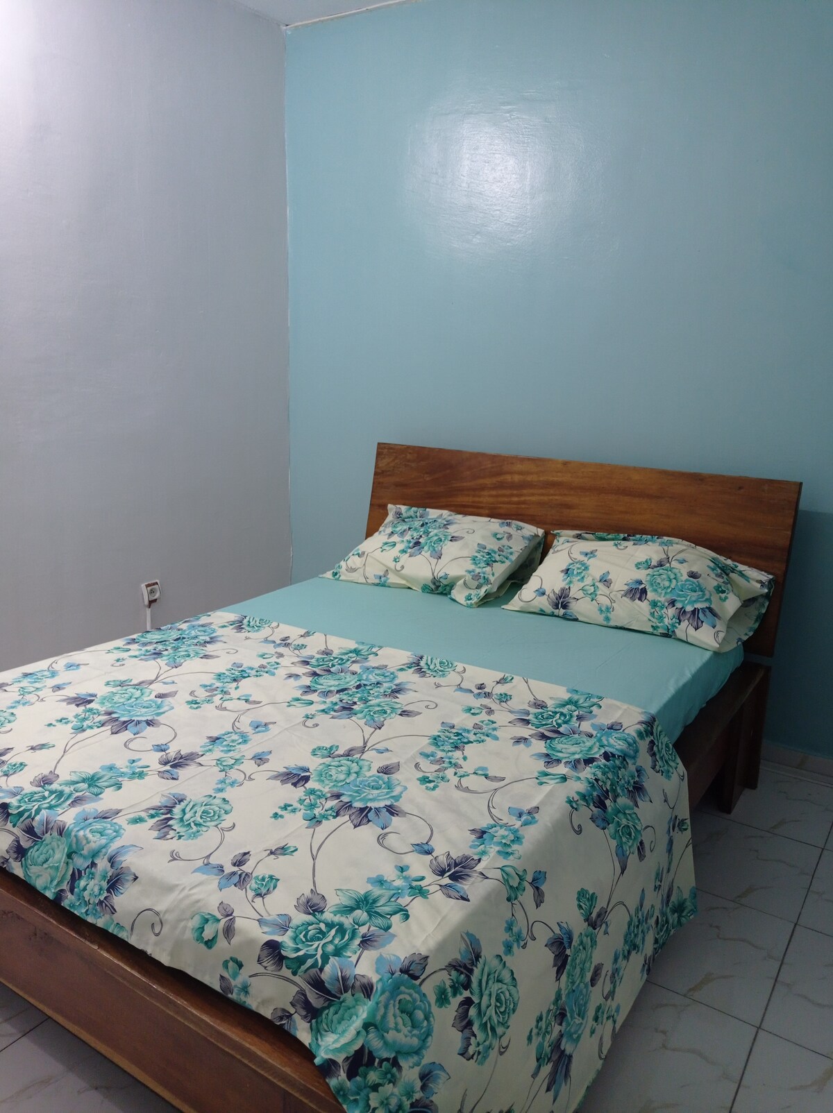 A serene bedroom features a wooden bed frame topped with a floral-patterned duvet cover and matching pillows. Soft blue walls contribute to a calming atmosphere, while tiled flooring completes the simple and clean aesthetic.
