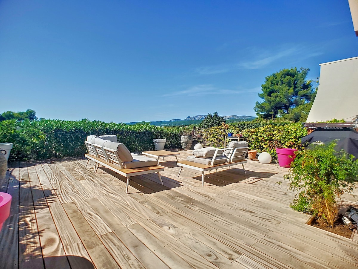 A spacious wooden terrace features comfortable lounge chairs set against a backdrop of greenery and distant hills. Potted plants add a touch of nature, while a barbecue area is visible, creating an inviting outdoor space for relaxation and enjoyment.