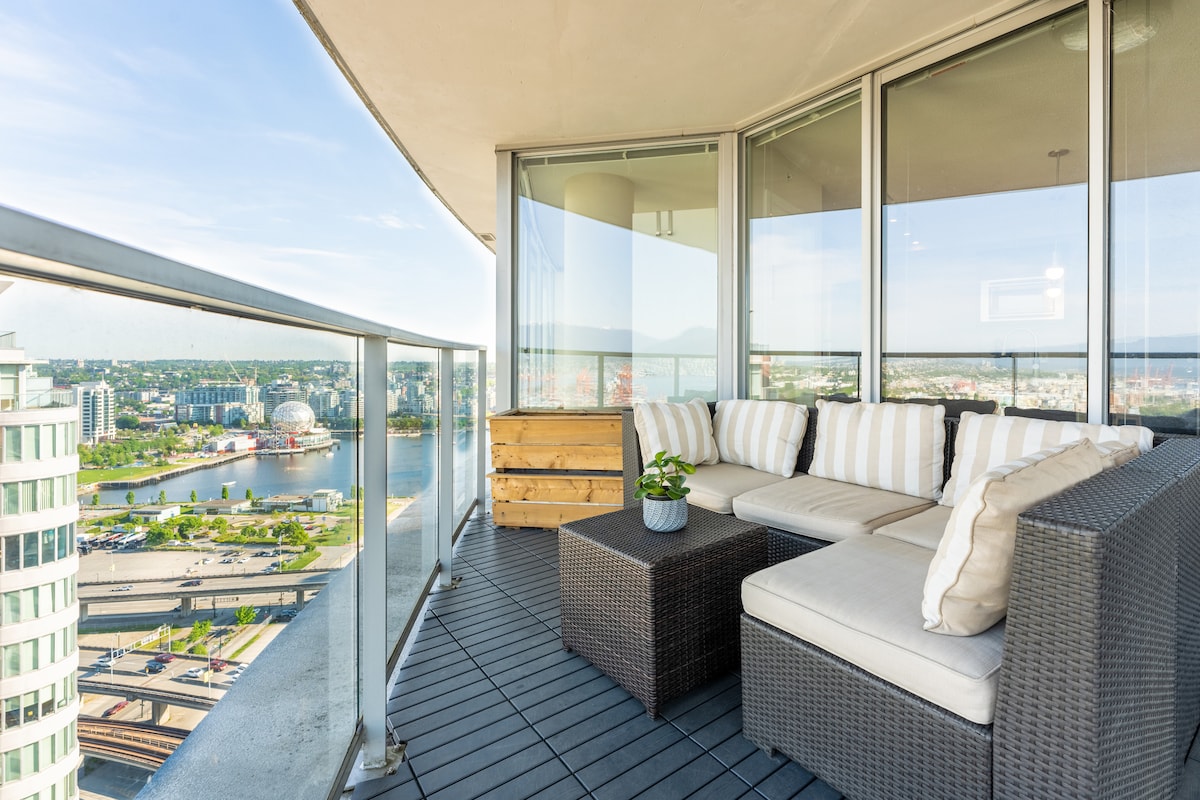 An oversized sundeck features a comfortable sectional sofa with striped cushions, positioned near a glass railing. A small table sits in front, offering a cozy space to relax while enjoying panoramic views of the cityscape and water below.