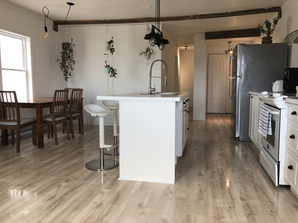 An open kitchen space features a central island with seating and wooden dining table nearby. Recessed lighting and wall-mounted plants add a touch of greenery. A modern refrigerator and stove are visible, complemented by a well-lit atmosphere created by large windows.