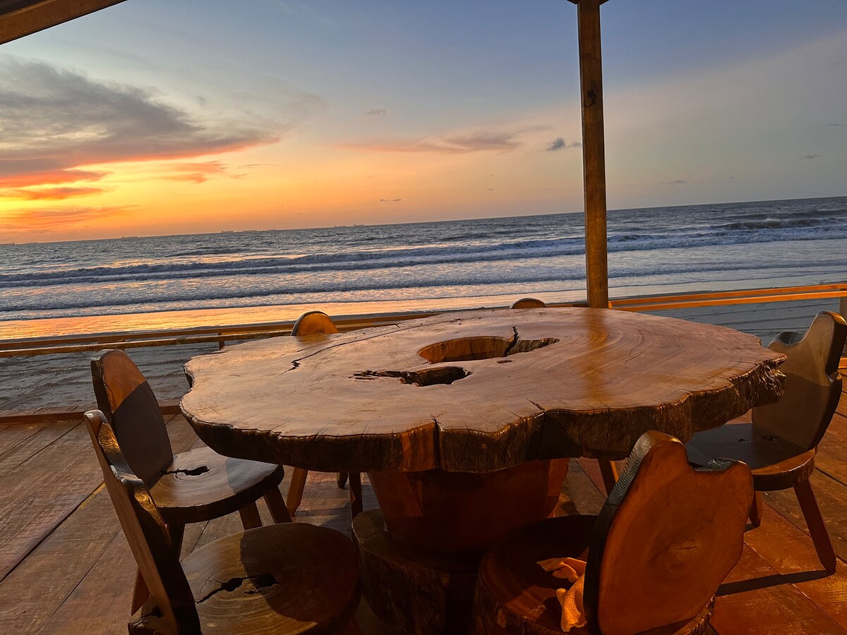 An outdoor dining area features a large wooden table with a natural edge and six circular chairs. The setting overlooks the ocean, with a vibrant sunset illuminating the sky and reflecting on the water's surface.