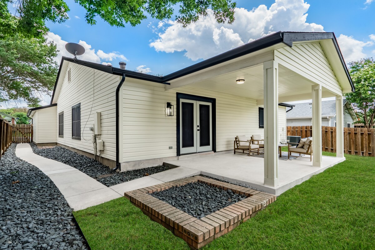 An inviting outdoor space features a covered porch with seating, surrounded by a well-maintained lawn. A pathway lined with decorative rocks leads to the entrance, while trees and a fence provide privacy and greenery in the background.