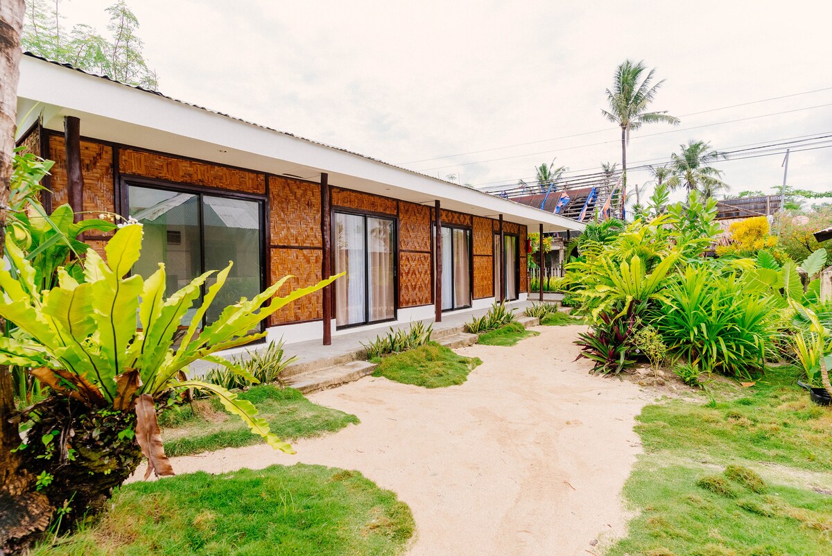 A serene exterior view of the hostel reveals a white structure with large windows framed by natural wood. Lush green plants and a sandy pathway are positioned around the building, contributing to a relaxed ambiance.