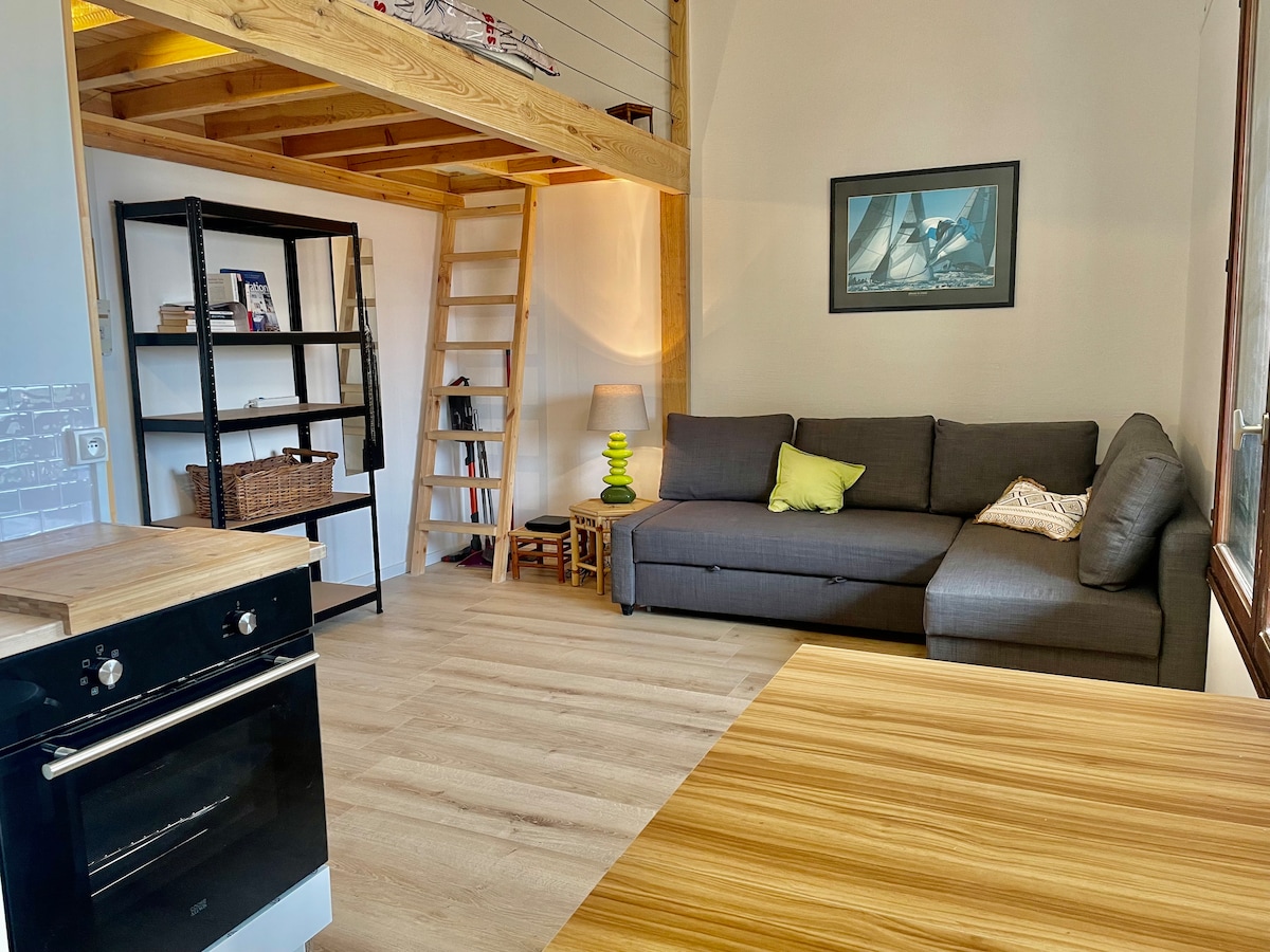 An open living area features a dark gray convertible sofa accompanied by a bright green cushion. A wooden ladder provides access to a mezzanine sleeping area. A wooden dining table is visible in the foreground, alongside an oven and a shelf holding books and decorative items.