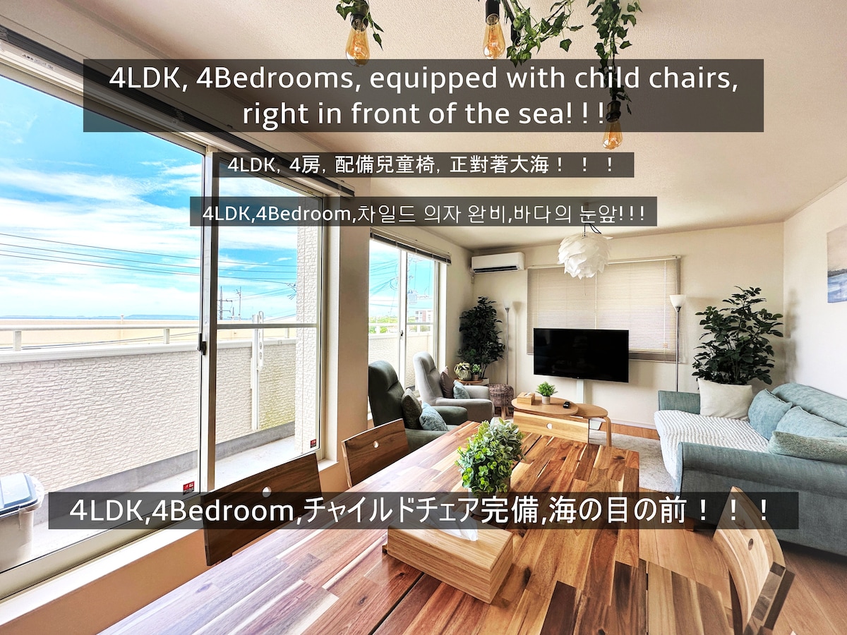 A bright living area features large windows that offer views of the nearby sea. Comfortable seating is arranged around a central table, and a child chair is included to accommodate families. The space is designed for relaxation and enjoying the coastal scenery.