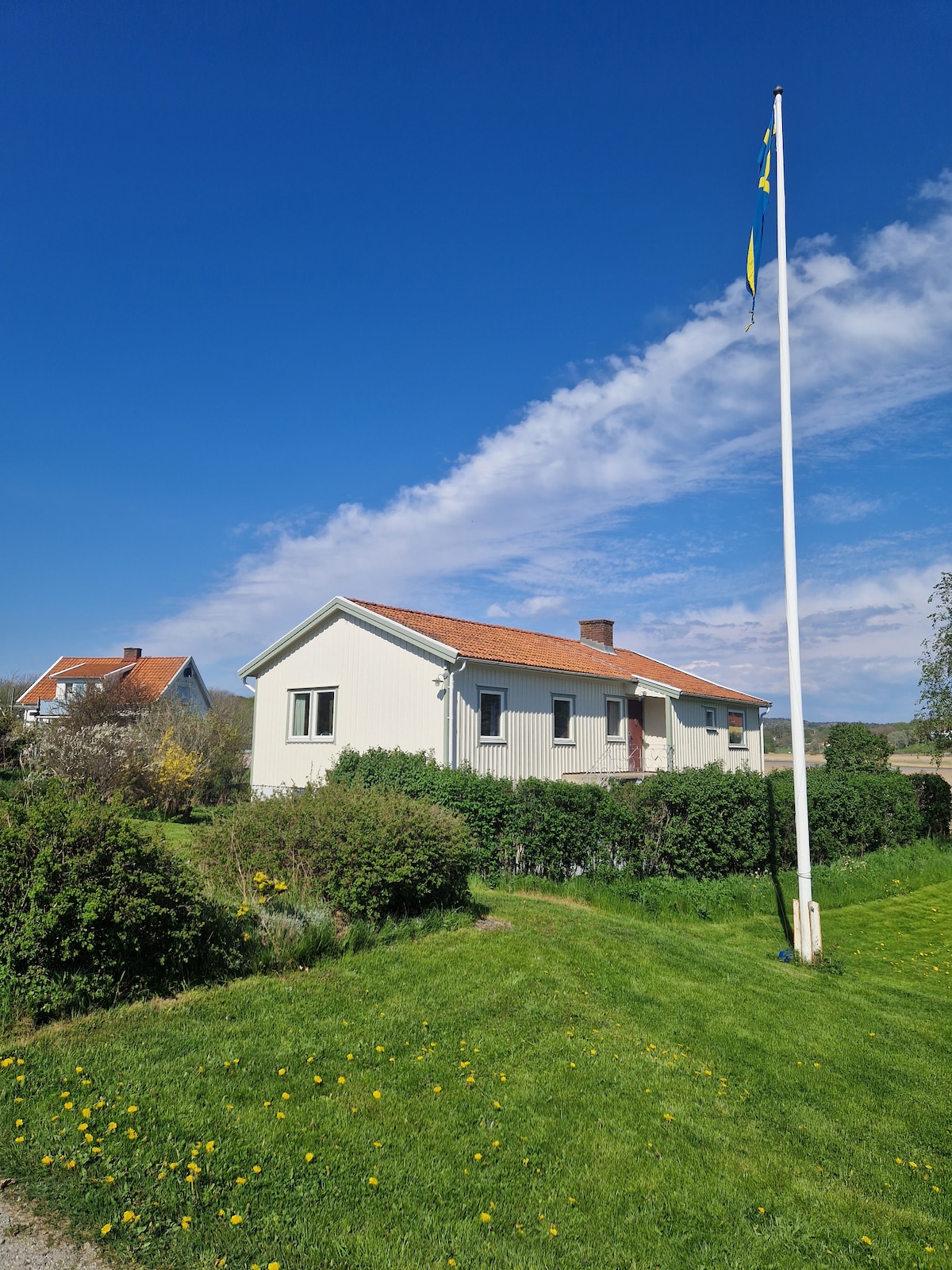 A single-story house with a light-colored exterior and a red-tiled roof stands surrounded by green grass and shrubs. A flagpole with the Swedish flag is prominently placed in the front yard, with a clear blue sky and wispy clouds in the background.