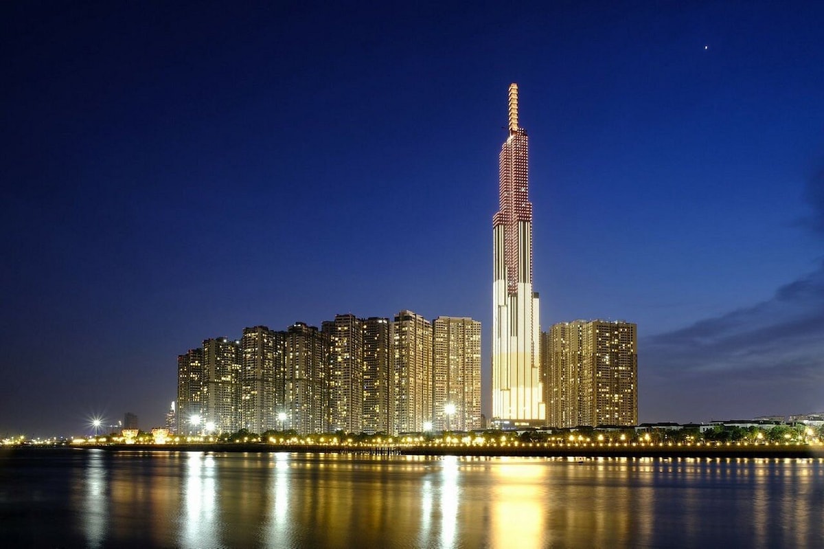 A modern skyscraper tower stands tall against a twilight sky, illuminated with dynamic lighting. Reflections shimmer on the calm water below, while surrounding buildings feature numerous lights, creating a lively cityscape atmosphere.