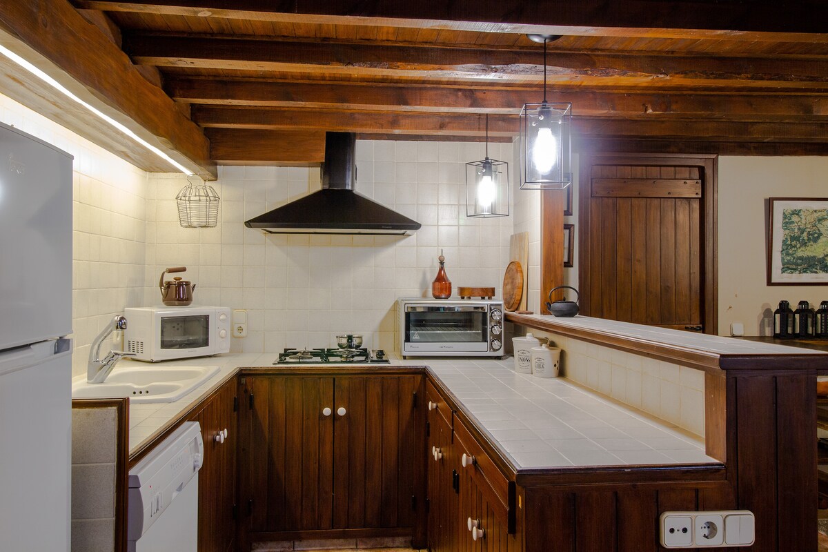 The kitchen features wooden cabinetry and exposed beam ceilings, creating a rustic ambiance. A gas stove and modern microwave are positioned on the countertop, alongside a sleek refrigerator. Soft lighting from hanging fixtures adds warmth to the space, enhancing its functionality.