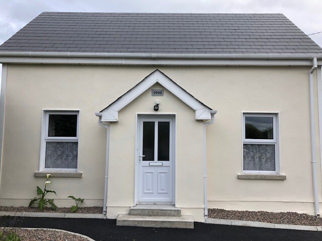 The exterior of a renovated cottage is displayed, featuring a light-coloured facade and a grey roof. Two windows flank a central white door, which is framed by a small porch. A neat pathway leads to the entrance, surrounded by gravel and greenery.