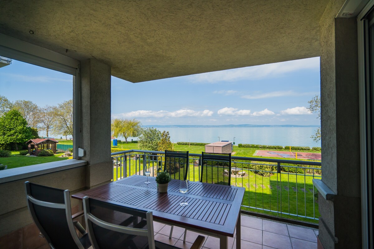 A spacious terrace is showcased, featuring a wooden dining table and chairs. The view extends to a lush green lawn leading to the serene lake, with clear skies above. The scene captures the inviting outdoor space, perfect for leisurely meals and enjoying the surrounding nature.