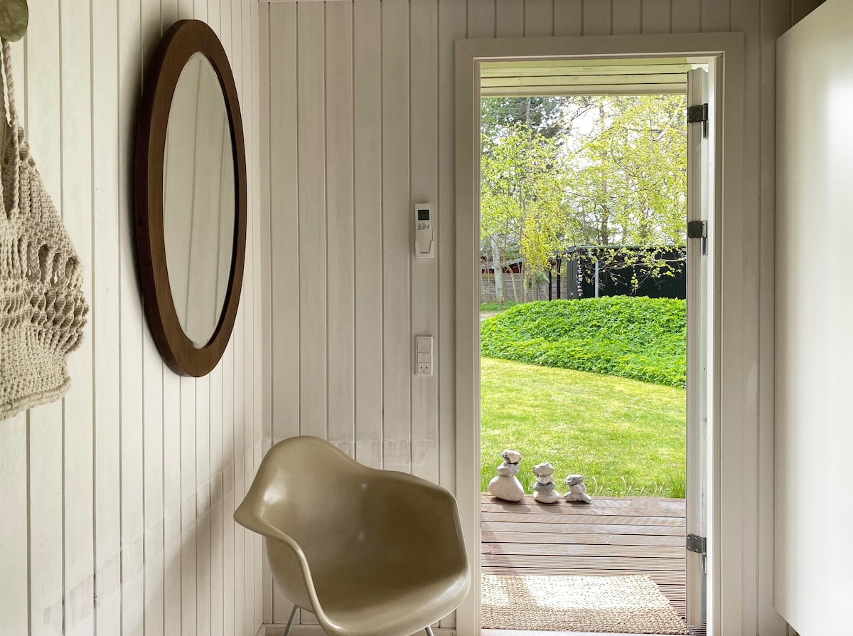 A light-filled entryway features a round mirror mounted on the white wooden wall. A sculptural chair is positioned beside the door, which opens to a grassy outdoor area. Smooth stones are arranged neatly on the wooden deck, inviting a sense of tranquility.