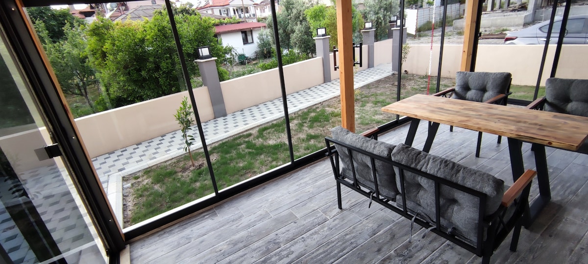 A spacious glass balcony area features a wooden dining table surrounded by four armchairs. The view includes a landscaped yard with greenery and a pathway. Natural light floods the space, highlighting the wooden flooring and the contemporary design.