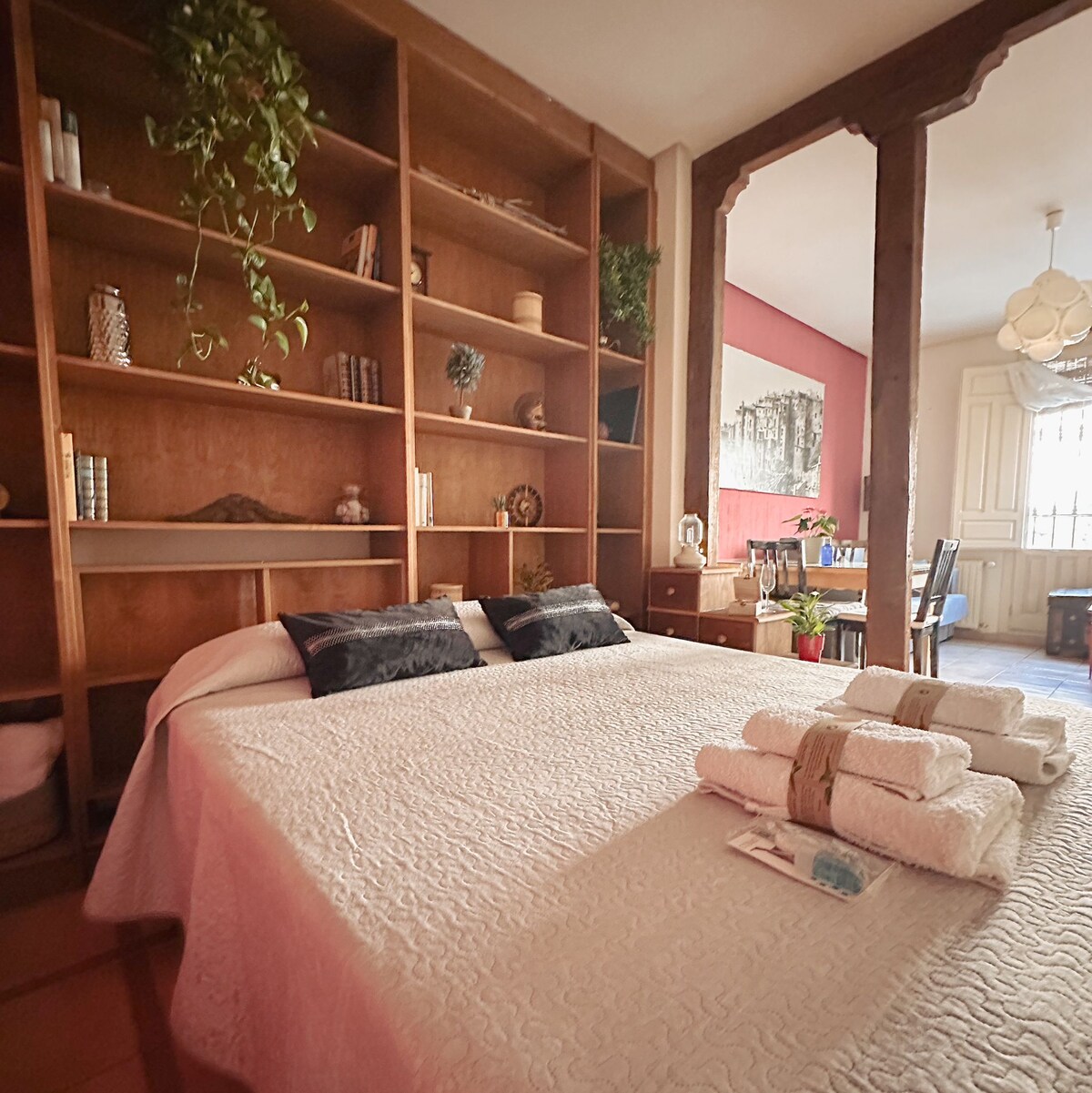 A spacious sleeping area features a neatly made bed with light bedding and two decorative pillows. A wooden bookshelf lines one wall, showcasing books and decorative items. A bright and inviting atmosphere is created by natural light filtering through the nearby window.