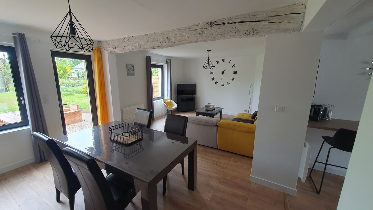 The open living area is highlighted by a dining table surrounded by four black chairs, with a cozy sofa and a yellow accent chair nearby. Large windows invite natural light, offering views of the garden. A modern clock adorns the wall, enhancing the contemporary decor.
