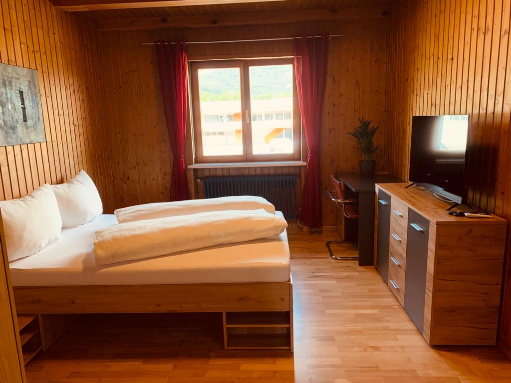 A cozy bedroom is presented with wood-paneled walls, featuring a spacious bed with crisp linens and two large pillows. A window allows natural light to enter, complemented by red curtains. A desk and a TV are positioned against the wall, providing functional amenities.